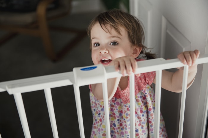 Vue de face d'une petite fille debout de l'autre côté d'une barrière de sécurité pour bébé. La petite fille s'accroche au haut de la barrière de sécurité et regarde la caméra par-dessus.