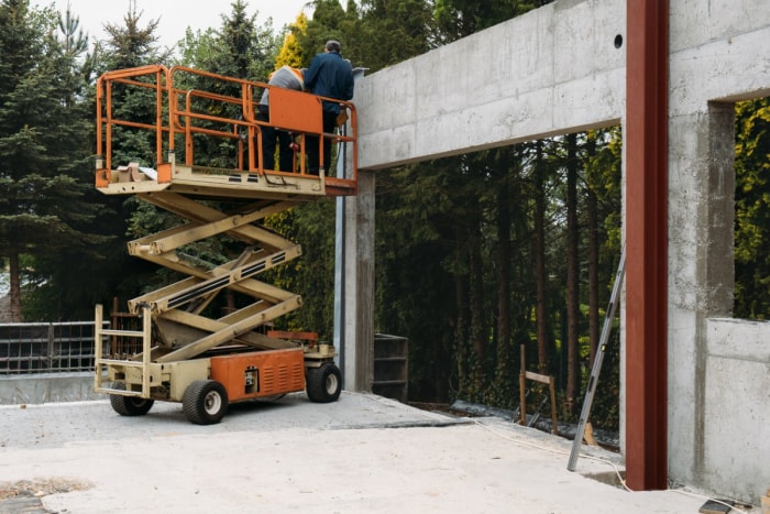 Une nacelle élévatrice à ciseaux orange en location est posée à côté d'un mur en béton.