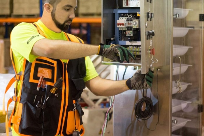 Une personne portant un gilet de sécurité noir et orange et des gants alors qu'elle travaille sur un boîtier électrique.