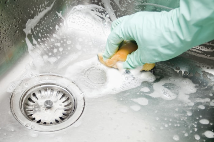 Des mains humaines avec des gants de protection récurent un grand évier de cuisine de forme carrée avec un tampon à récurer, des bulles de savon.