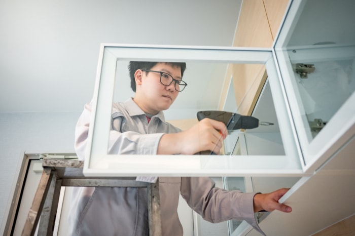 Un homme asiatique sur une échelle utilise une perceuse pour installer une nouvelle porte d'armoire de cuisine transparente.