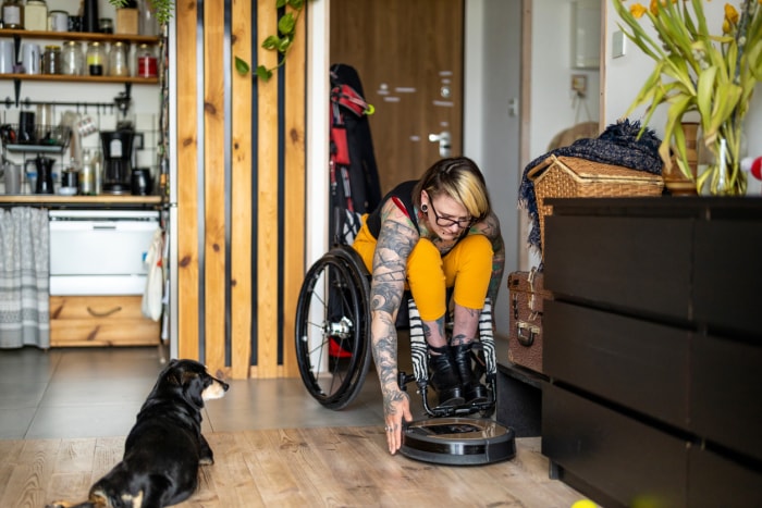 iStock-1396837798 Remboursement d'impôt Améliorations domiciliaires Une femme en fauteuil roulant ajuste un robot nettoyeur intelligent