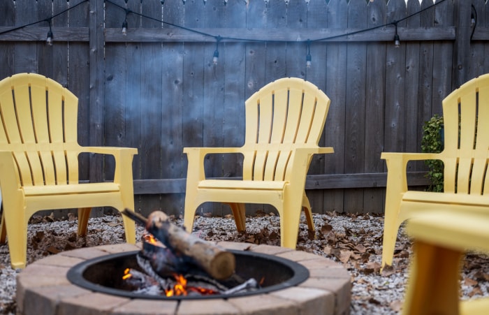 Un foyer avec des bûches, entouré de chaises en plastique jaune, dans l'arrière-cour recouverte de feuilles mortes et entourée d'une clôture en bois.