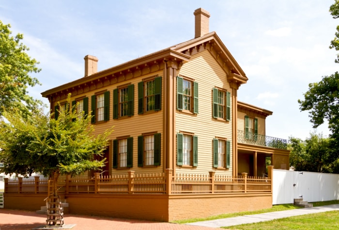 une maison de style néo-grec avec un extérieur beige et des volets vert foncé, située sur un terrain d'angle avec un arbre, est la maison d'Abraham Lincoln