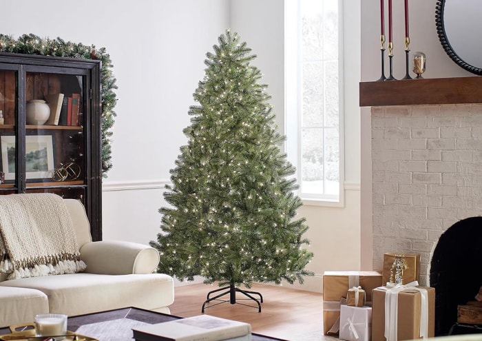 Le sapin de Noël National Tree Company Full Downswept dans un salon neutre à côté d'une cheminée en briques avec des cadeaux emballés.