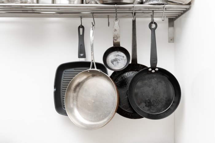 Différentes casseroles de différentes tailles et formes pour cuisiner et frire, suspendues à des crochets métalliques sur une étagère de cuisine avec un mur blanc en arrière-plan