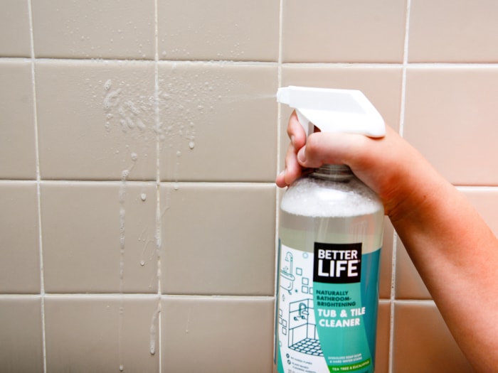 Une personne pulvérisant le nettoyant pour baignoire et carrelage Better Life sur le carrelage de la douche.