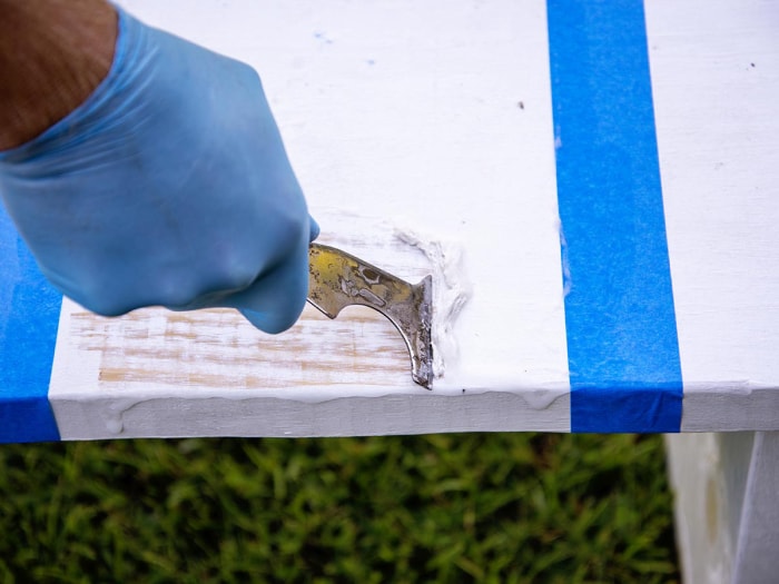 Une main gantée utilise un outil de grattage pour décaper la peinture d'une table.