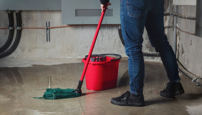 Un homme nettoie un sous-sol inondé avec un seau rouge et une vadrouille.