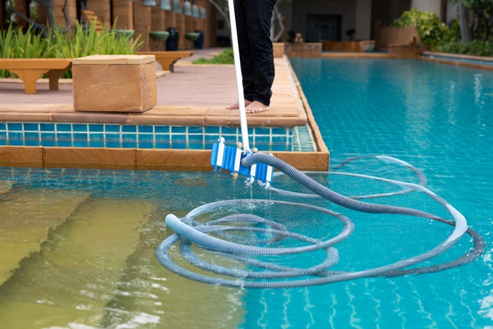 Une personne dont seules les jambes sont visibles nettoie une piscine de jardin à l'aide d'une brosse.