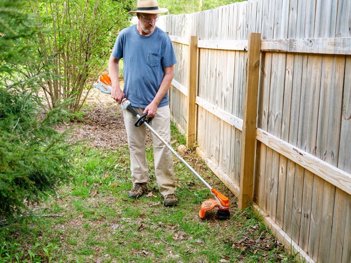 Personne en chemise bleue utilisant un coupe-herbe orange pour couper l'herbe le long d'une clôture en bois