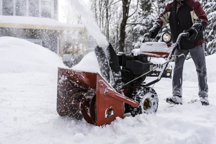 homme utilisant une souffleuse à neige devant une maison couverte de neige et un aménagement paysager