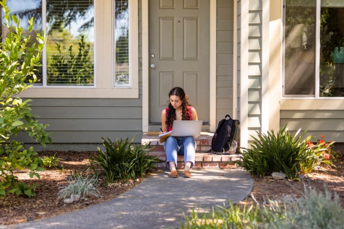 Une femme est assise devant une maison grise, assise devant un ordinateur portable, écrivant dans un cahier. 