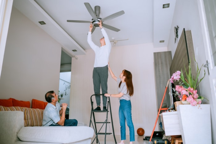 Un homme aide sa famille à changer la direction du ventilateur de plafond sur une échelle.