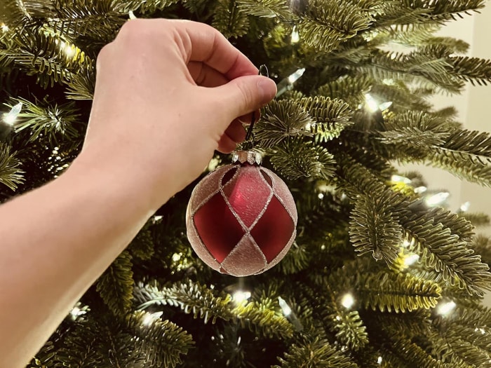 Une personne accroche une décoration de Noël rouge et or sur les branches pré-éclairées de l'arbre à bascule de Balsam Hill.