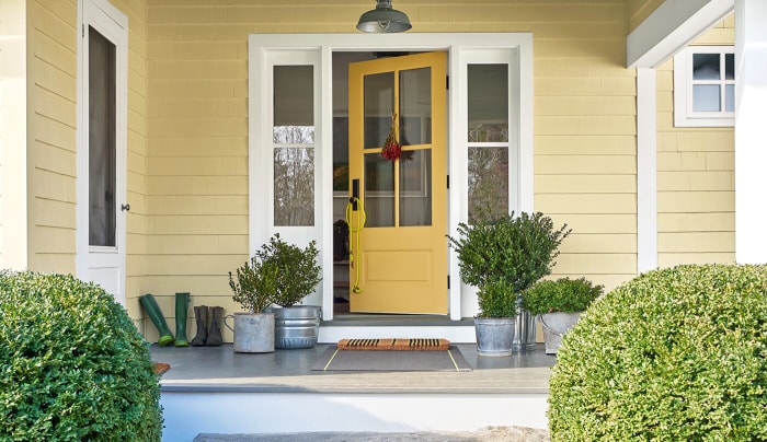 Deux plantes vertes en pot se trouvent à côté d'une porte jaune sur le porche d'une maison à bardage jaune.