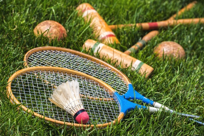 Une paire de raquettes de badminton en bois et un volant en plumes reposant dans l'herbe, ainsi qu'une paire de maillets et de boules de croquet en arrière-plan. Des jeux familiaux à l'ancienne pour tous les âges.