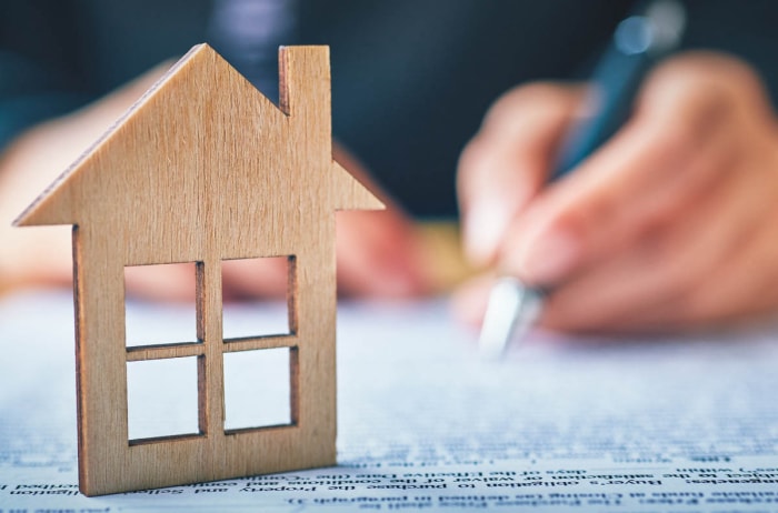 Une petite maquette de façade de maison en bois est posée sur une table avec une personne qui écrit en arrière-plan.