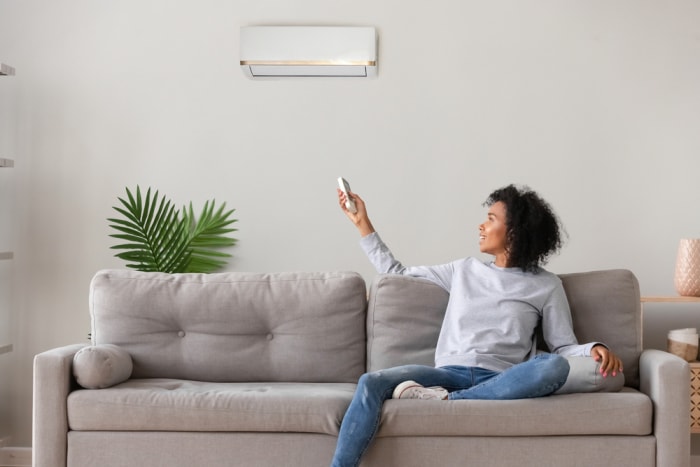 Une femme est assise sur un canapé et utilise une télécommande pour contrôler un climatiseur monté au-dessus de sa tête. 