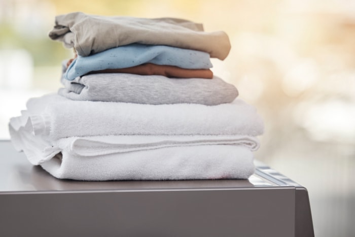 Une pile de serviettes blanches pliées et de t-shirts neutres posés sur une machine à laver.