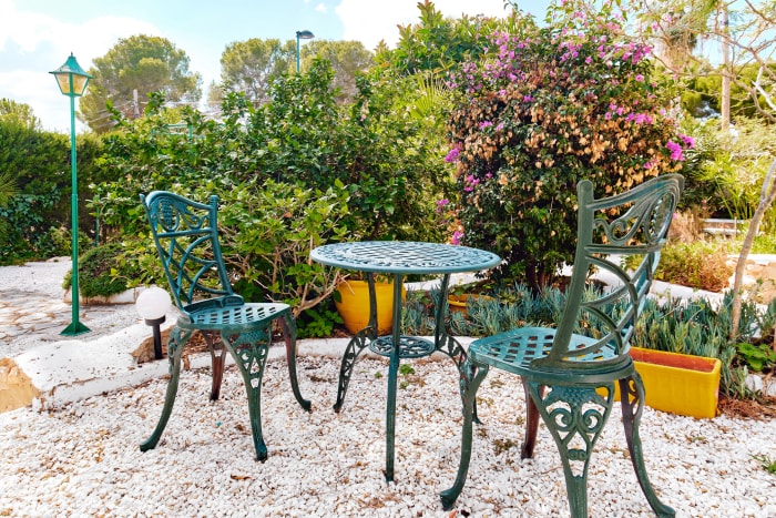 Chaises et table en métal de couleur sarcelle sur une terrasse extérieure avec des fleurs en fleurs en arrière-plan.