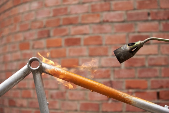 Utilisation d'un chalumeau pour enlever la peinture orange sur un vélo en métal.