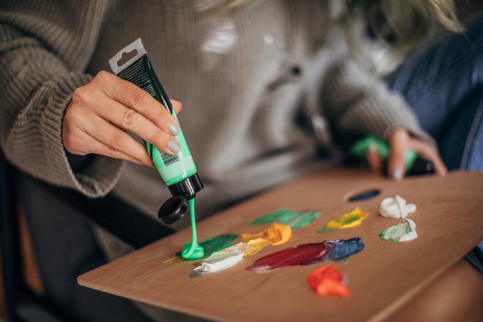 Un tube de peinture acrylique vert clair est pressé sur une palette par une femme portant un pull.