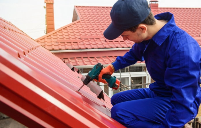 Un homme portant une casquette bleue et une combinaison de travail bleue utilise une perceuse pour installer un toit rouge. 