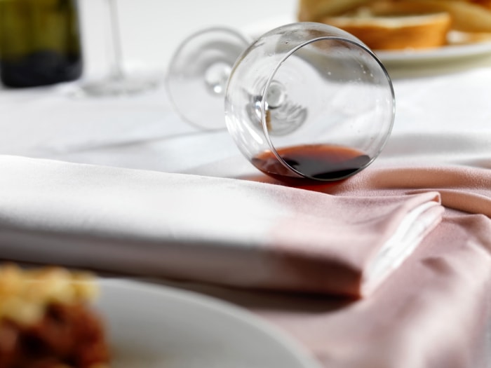 Un couvert sur une table avec une nappe blanche et une serviette avec un verre de vin rouge renversé et tachant la nappe.