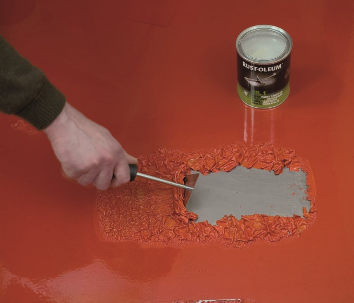 Une main utilisant un outil pour décaper de la peinture rouge sur du métal.