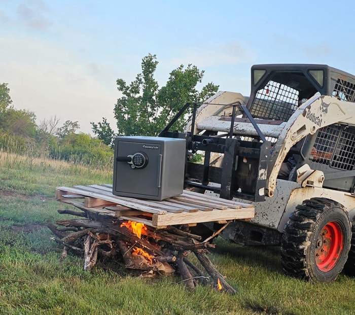 Le coffre-fort domestique SentrySafe est posé sur une palette maintenue au-dessus d'un feu de joie par un skid steer.