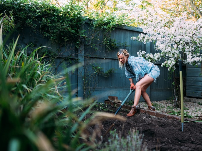 Une femme mature portant des bottes et des gants en cuir creuse un grand parterre de jardin dans son jardin.