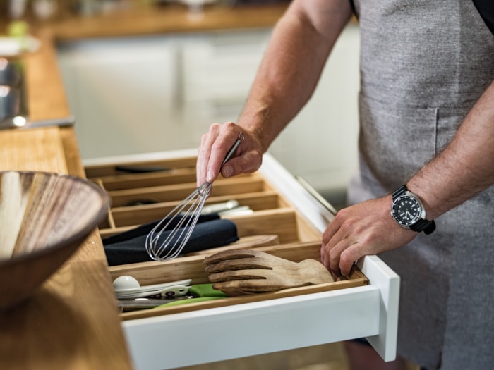 Mains d'un homme prenant le fouet métallique dans le tiroir à ustensiles d'une cuisine familiale. Il porte des vêtements décontractés avec un tablier de chef.