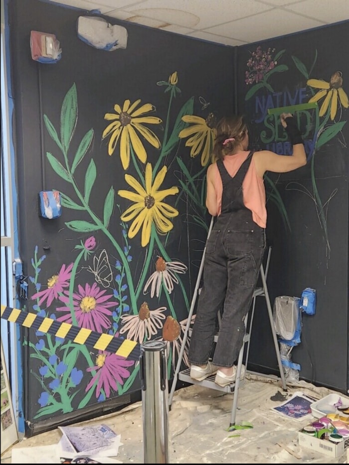 Une personne se tient sur un escabeau et peint une fresque florale colorée sur un mur noir.