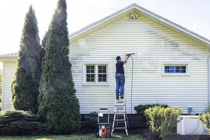 Un homme mature se tient sur une échelle et utilise un nettoyeur haute pression pour nettoyer le revêtement de la maison.