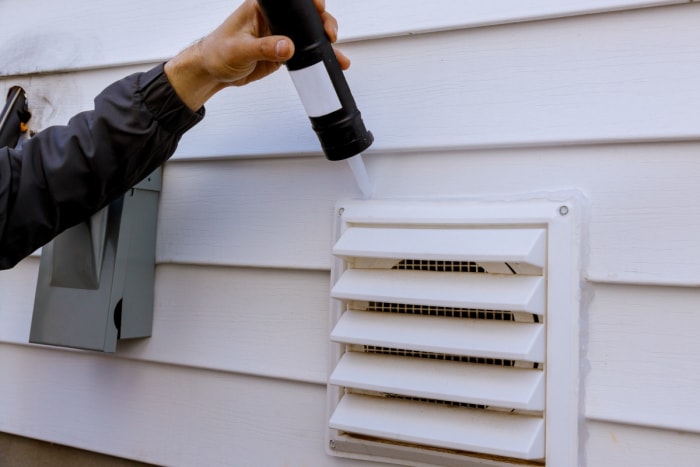 Le bras d'une personne est vu en train d'utiliser du calfeutrage pour sceller la ventilation extérieure d'une maison.