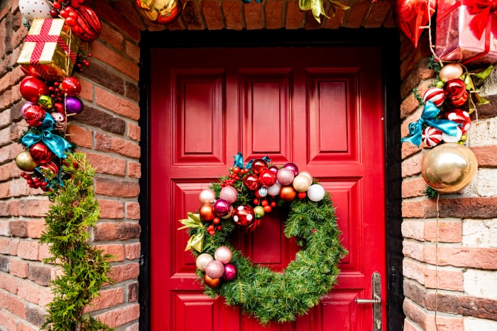 En attendant Noël - Porte d'entrée en bois rouge décorée de couronnes, guirlandes, nains, cadeaux,...