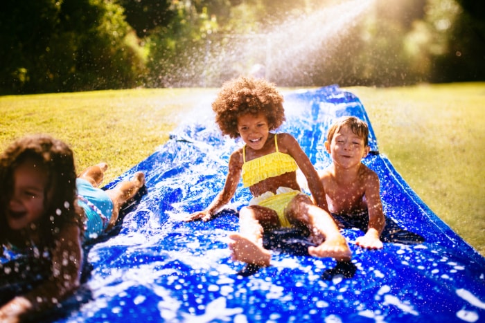 Fille noire rieuse et ses amis jouant ensemble sur un toboggan aquatique dans leur jardin au soleil.