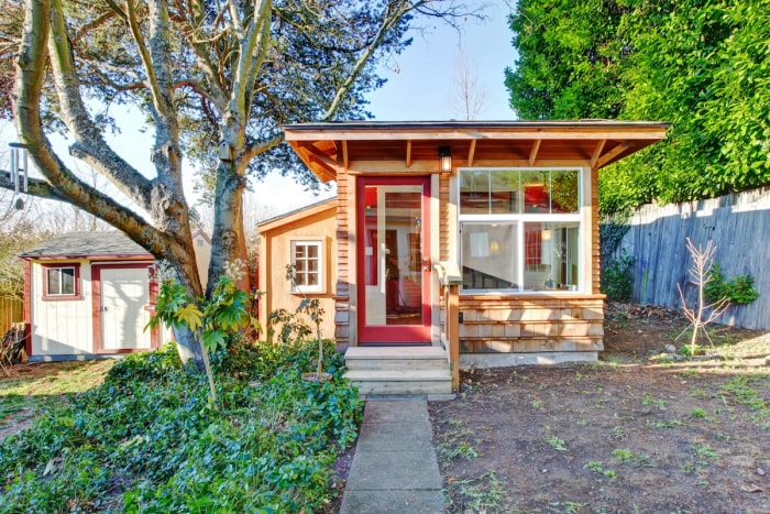 Petite maison en bois clair avec fenêtres et porte d'entrée rouge.