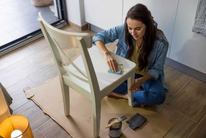 Une jeune femme aux longs cheveux bruns ponce le dessus d'une chaise en bois blanc avec du papier de verre, assise par terre à l'intérieur pendant la journée, écoutant de la musique avec ses écouteurs blancs, un pot de peinture avec un pinceau posé sur le sol à côté de la chaise, la cuisine en arrière-plan