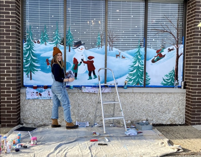 Une femme se tient à côté d'une peinture murale qu'elle peint d'une scène enneigée à l'extérieur sur une fenêtre.