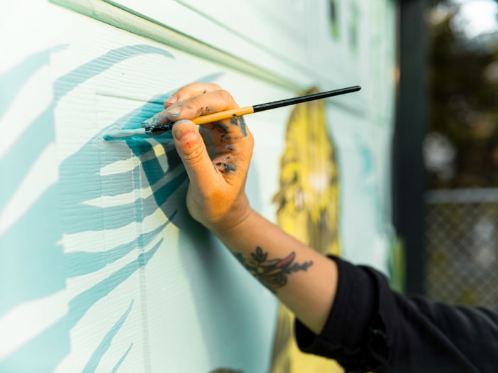 Une main avec des traces de peinture dessus tient un pinceau et ajoute des lignes bleu sarcelle à une peinture murale.