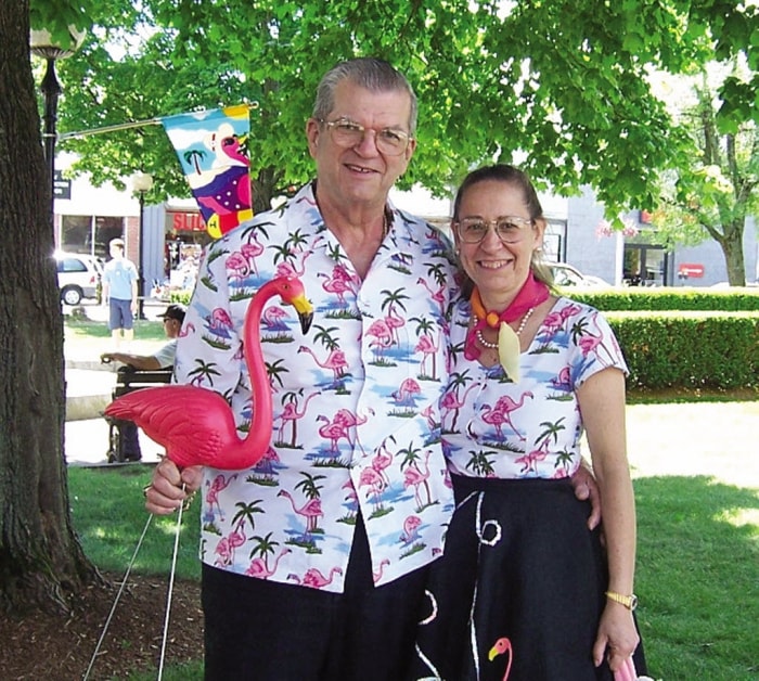 un homme et une femme âgés portant des chemises de flamants roses assorties se tiennent dehors sous un arbre tenant un flamant rose en plastique