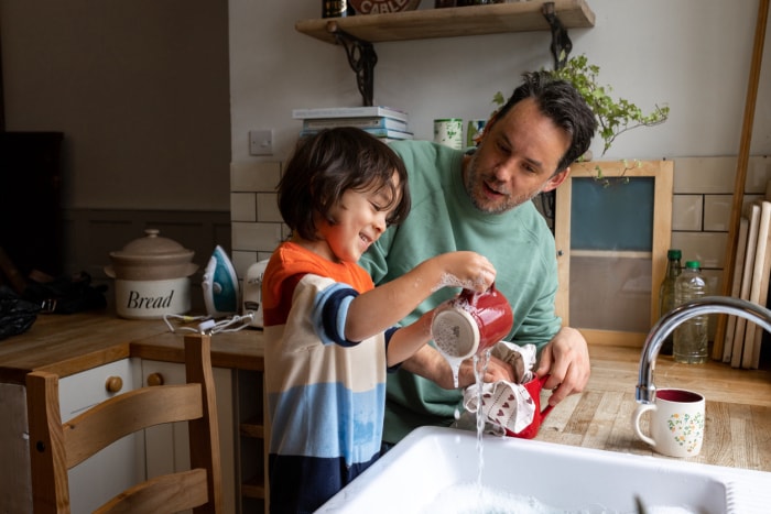 Plan à la taille d'un enfant de sexe masculin et de son père en train de laver et d'essuyer la vaisselle dans leur maison. Ils sont tous deux souriants et portent des vêtements décontractés.