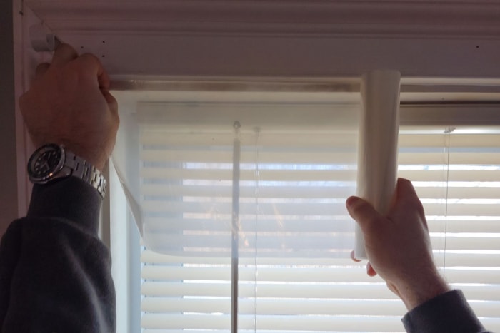 Une personne déroulant le meilleur kit d’isolation de fenêtre sur une fenêtre avec des mini-stores blancs.