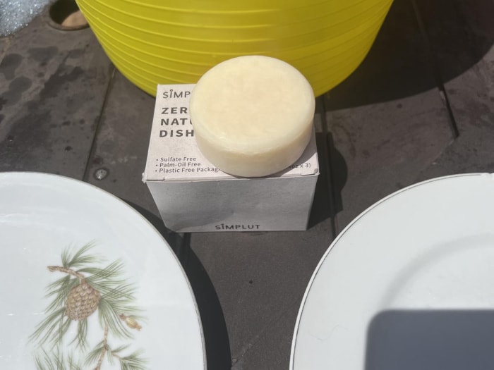 Barre de savon à vaisselle Simplut à côté de deux assiettes blanches avec des pommes de pin dessus.