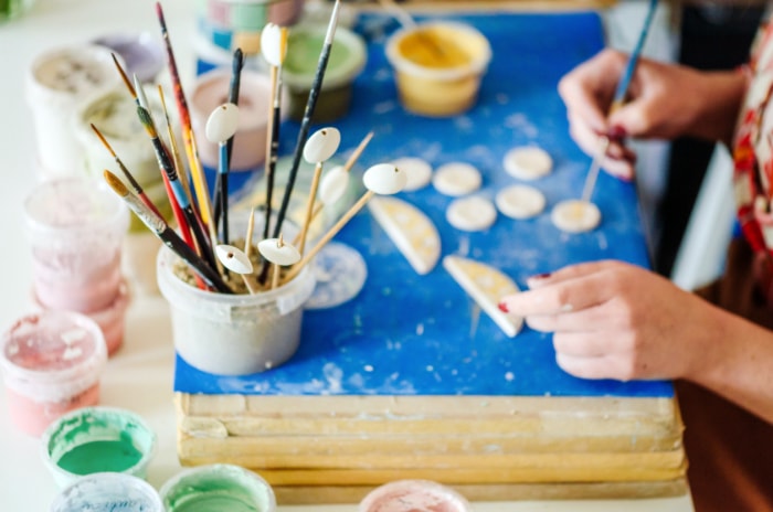 Une femme peint de petits objets en céramique à son poste de peinture installé avec différentes peintures et pinceaux.