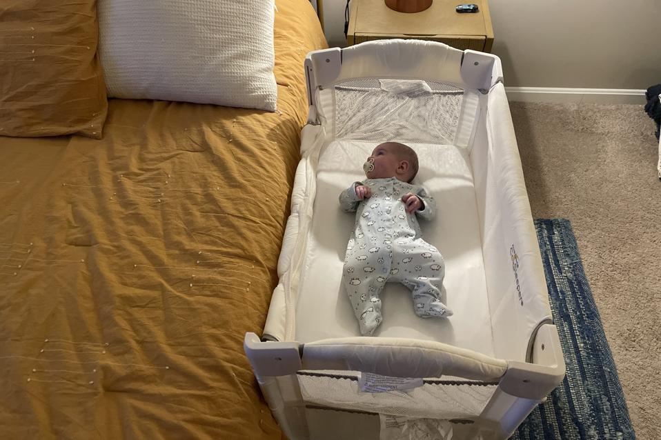 A baby lying in the Arm’s Reach Mini Ezee 2-in-1 Co-Sleeper Bassinet next to a bed.