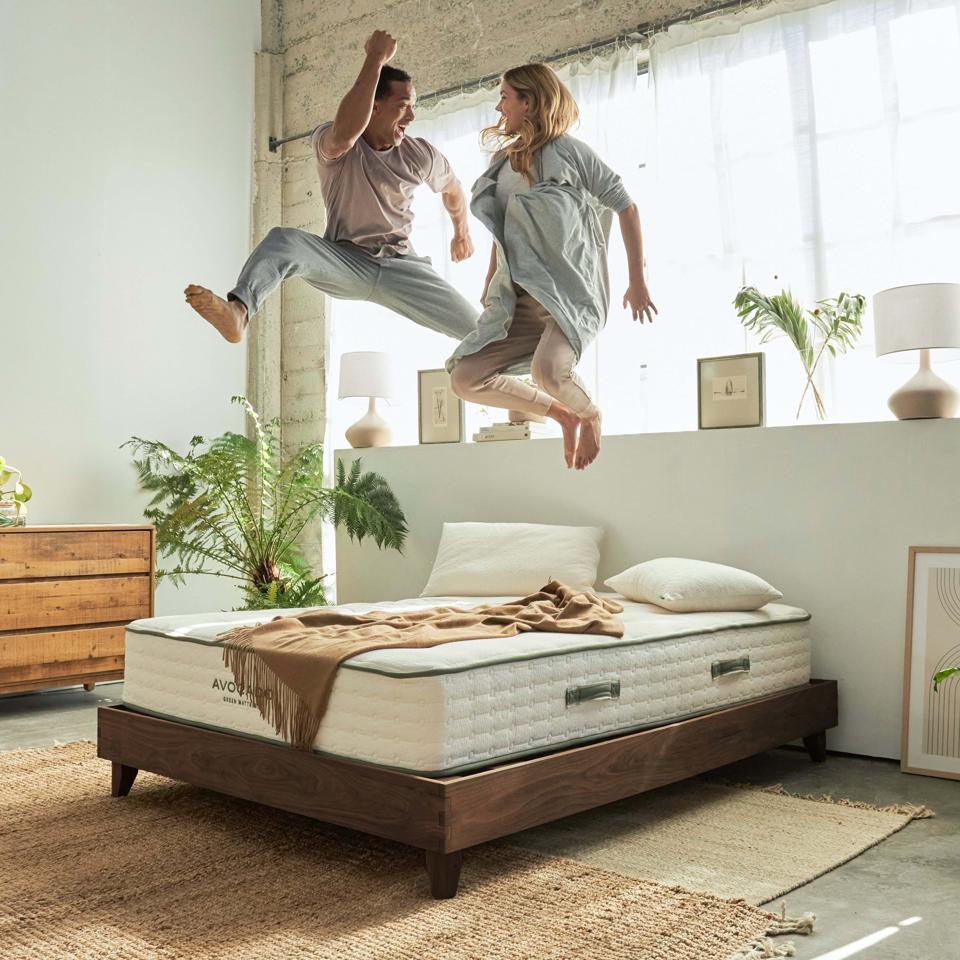 Two people jumping on an Avocado mattress and Avocado bed frame in bright bedroom.