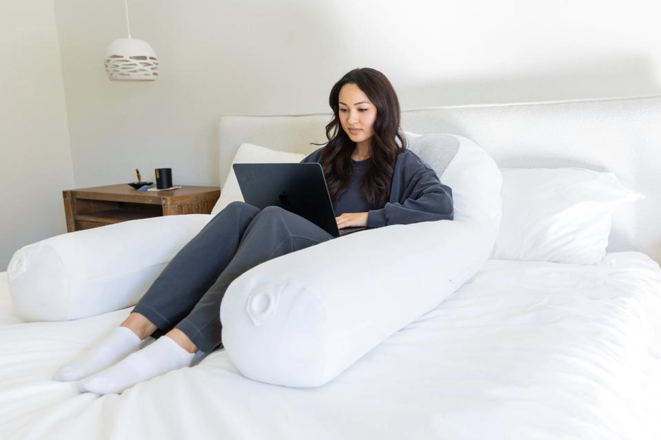 Woman sitting upright against a white U-shaped body pillow on a bed, with laptop in lap.